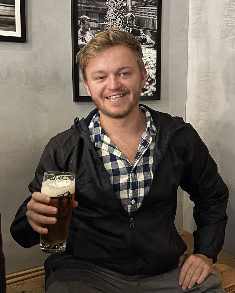 Brett holding a glass of beer with a beer mug logo on the glass, in a casual setting